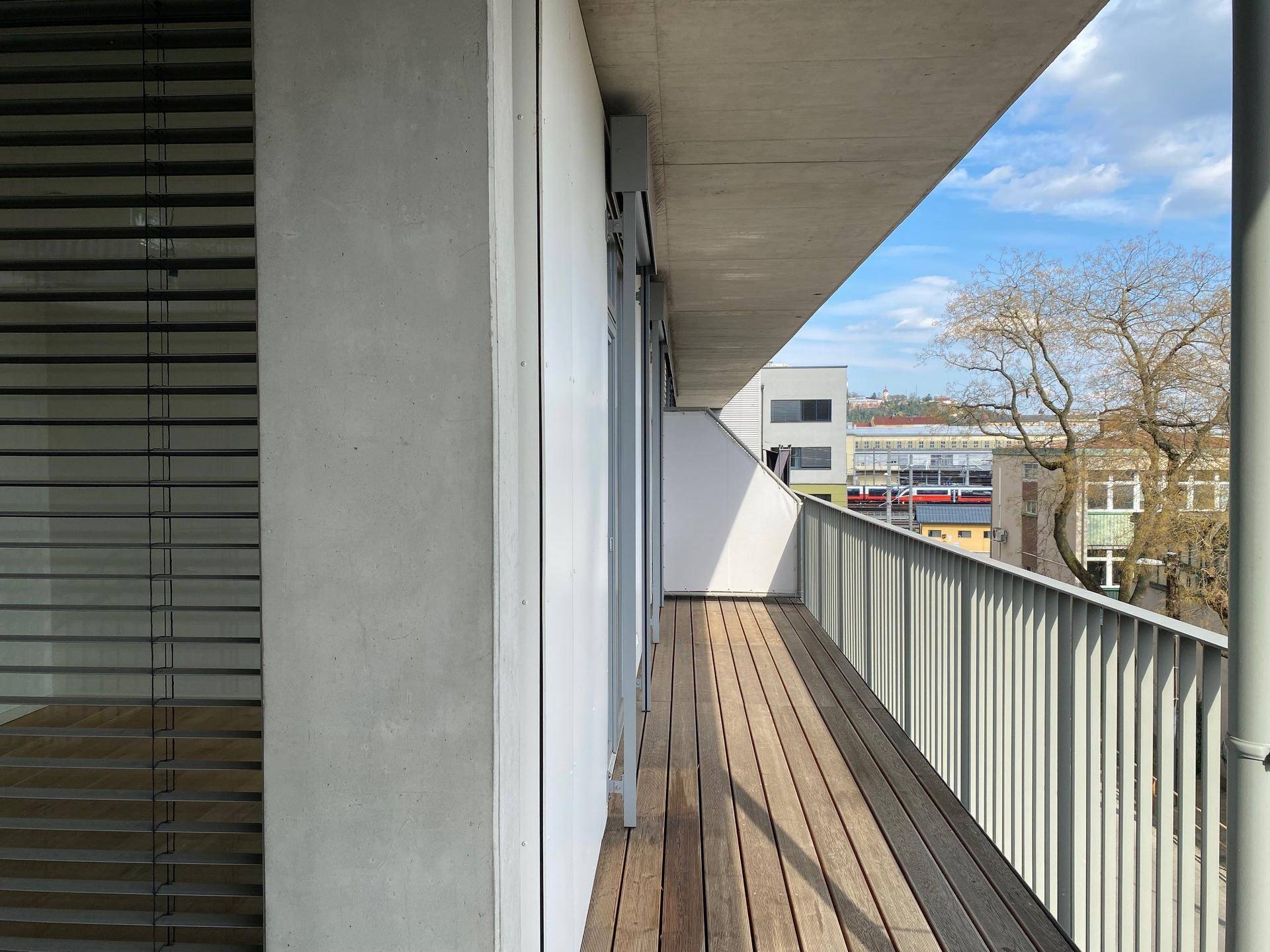 Langer Balkon mit Holzbelag und Metallgeländer, bietet einen seitlichen Blick auf die Stadt.