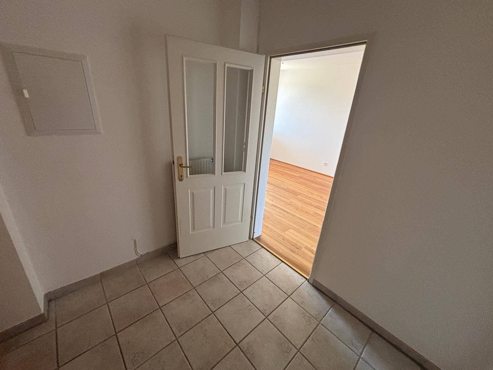 Flur mit Fliesenboden und offener Tür zu einem Zimmer mit Holzparkett.