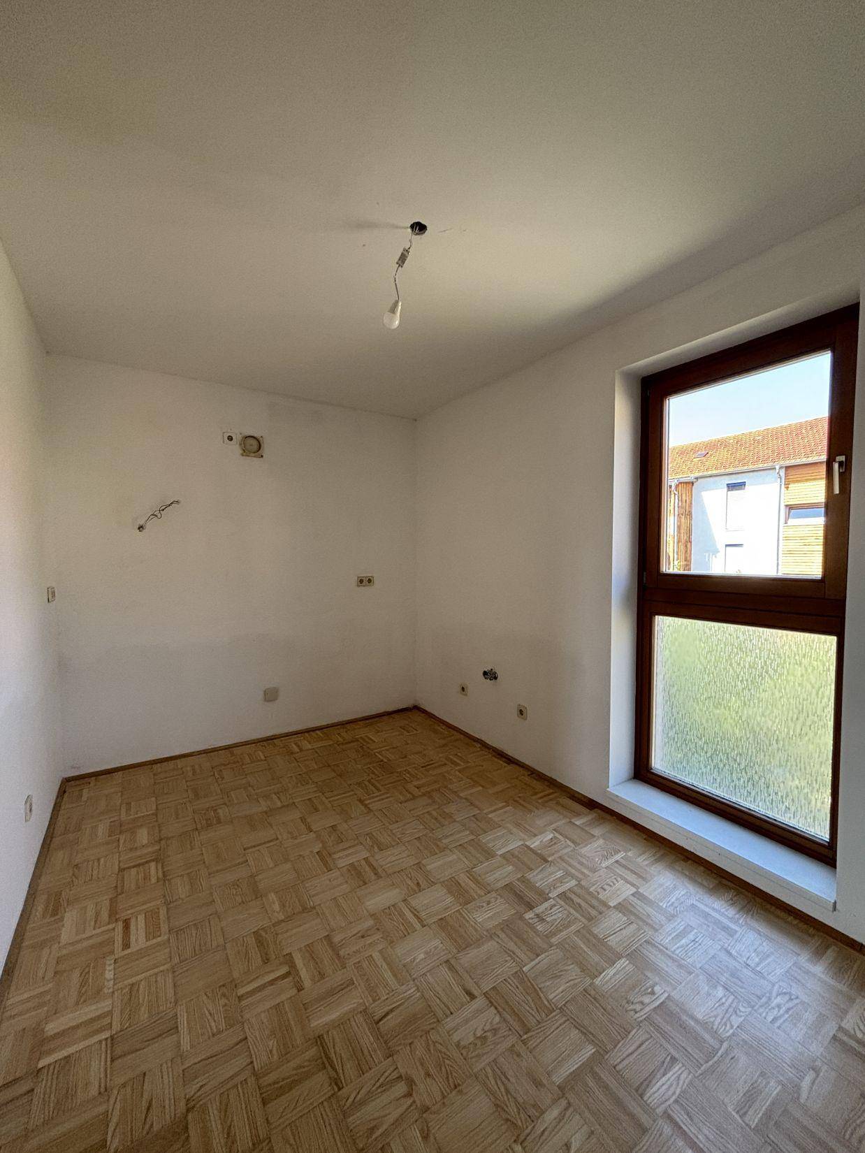 Leeres Zimmer mit Parkettboden und einem großen Fenster, das viel Tageslicht hereinlässt.