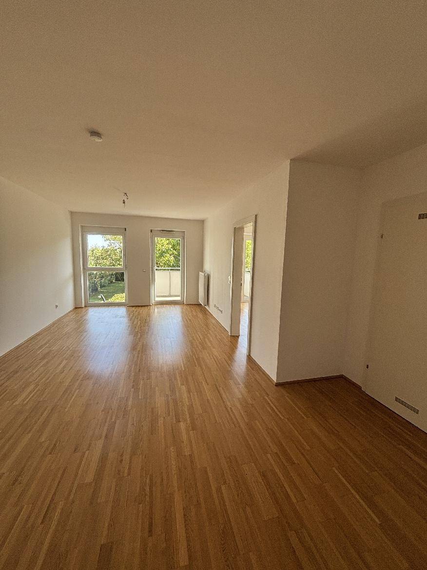 Geräumiges Wohnzimmer mit hellem Parkettboden und Zugang zu einem Balkon, lichtdurchflutet.