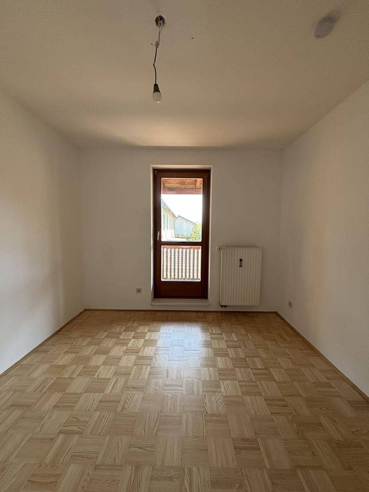 Leeres Zimmer mit Parkettboden, Heizkörper und Zugang zu einem Balkon.