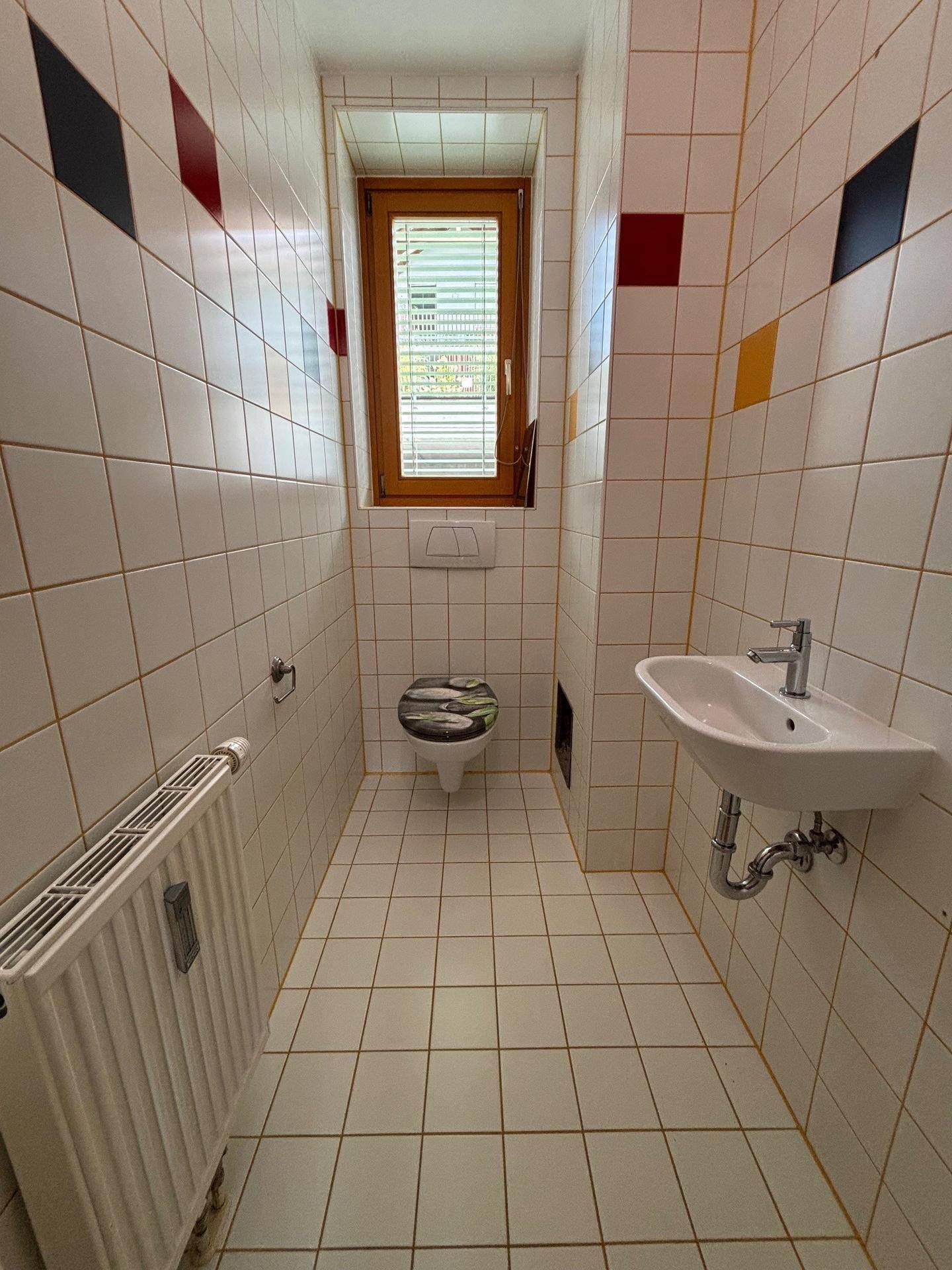 Gäste-WC mit Toilette, Waschbecken, Fenster und weißen Fliesen mit farbigen Akzenten.