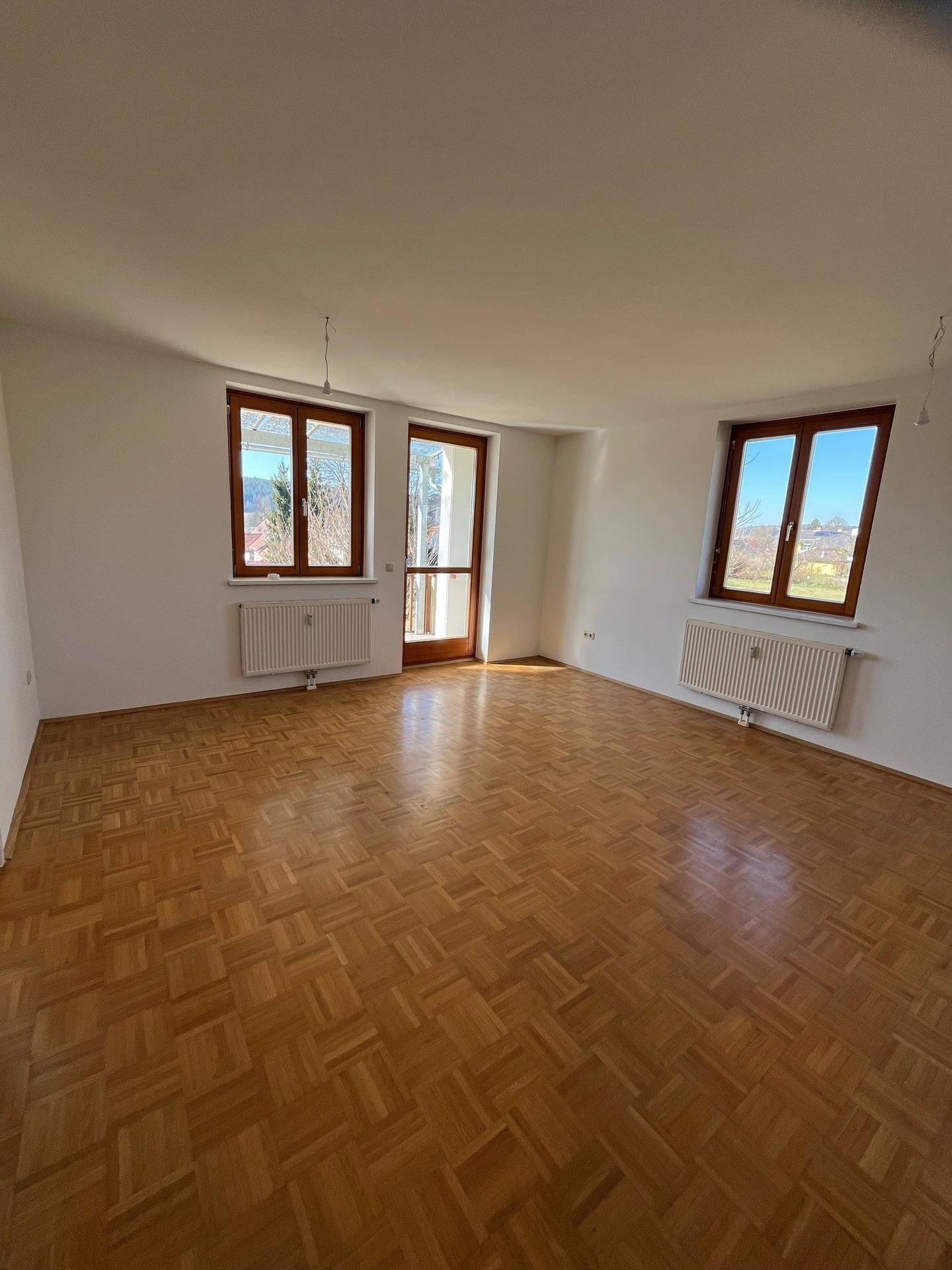 Geräumiges Wohnzimmer mit Parkettboden, mehreren Fenstern und Zugang zu einem sonnigen Balkonbereich.
