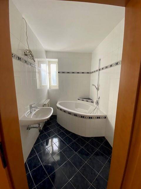 Badezimmer mit Eckbadewanne, Waschbecken, blauen Fliesen und einem Fenster für Tageslicht.