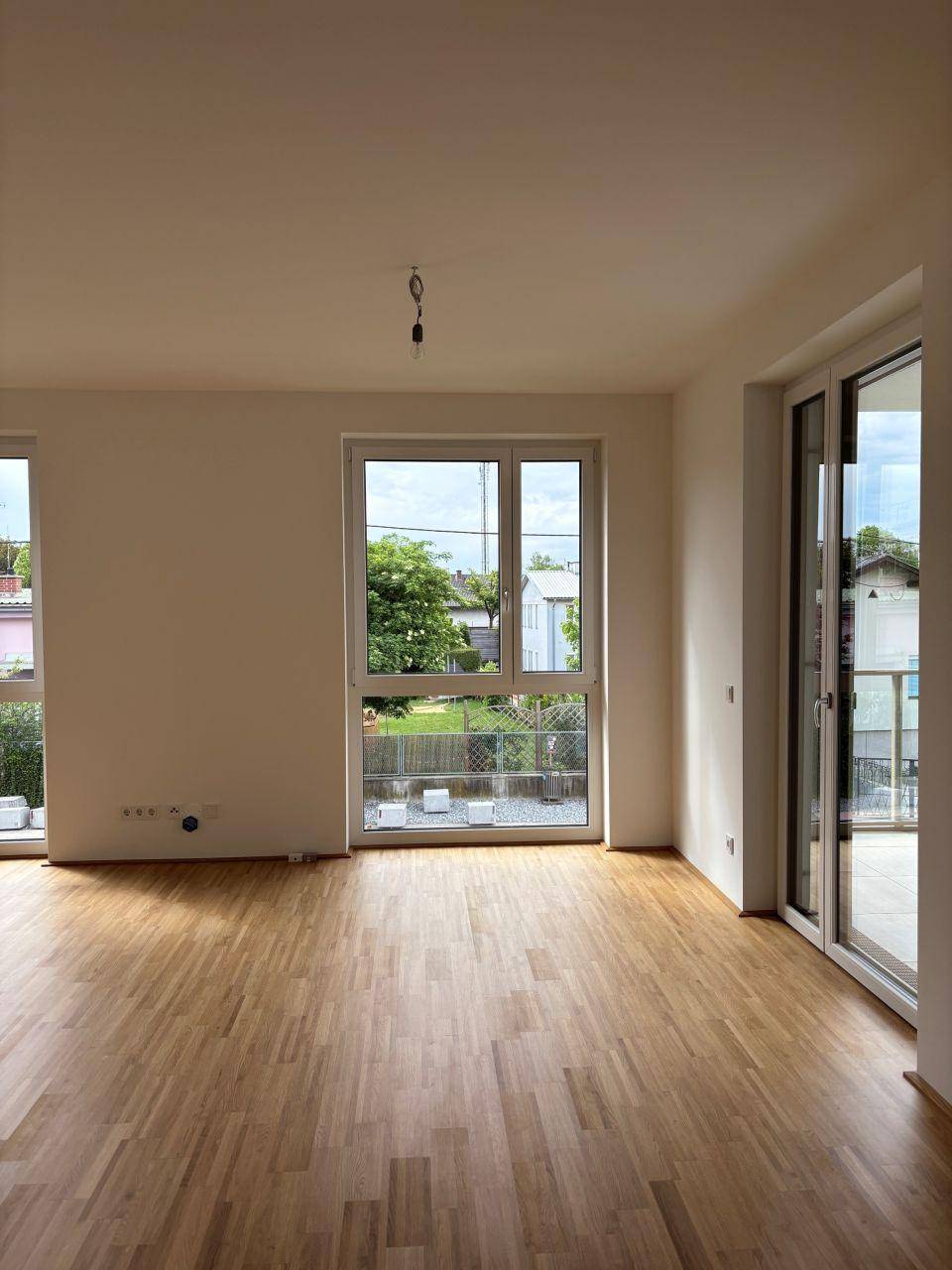 Leeres Wohnzimmer mit hellem Parkettboden und großen Fenstern zum Garten.