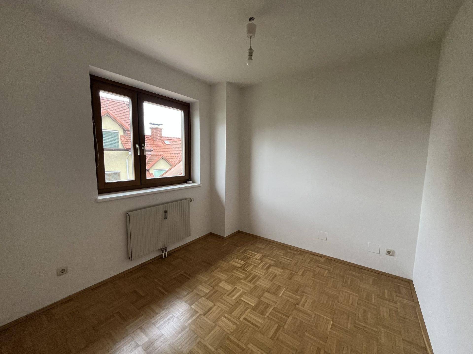 Helles Schlafzimmer mit Parkettboden, weiß gestrichenen Wänden und Blick auf die Dächer.