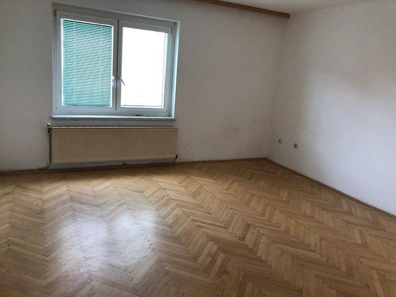 Leeres Zimmer mit Fischgrätparkett, großem Fenster und Heizkörper.