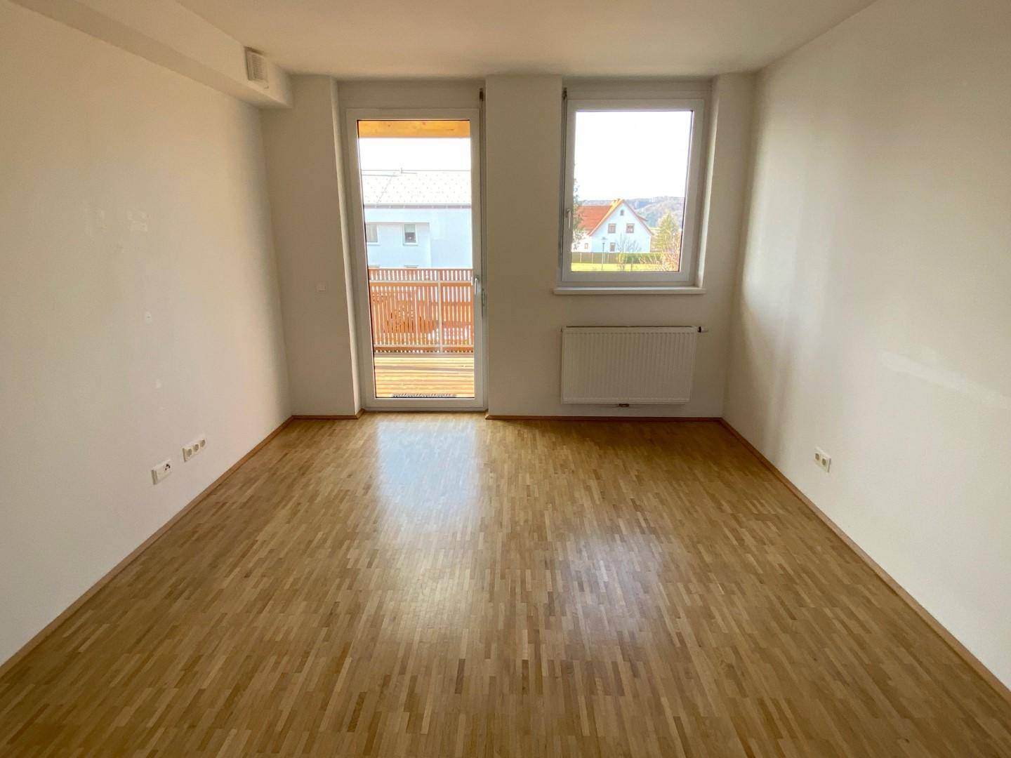 Helles Zimmer mit Parkettboden, großem Fenster und Zugang zu einem Balkon.