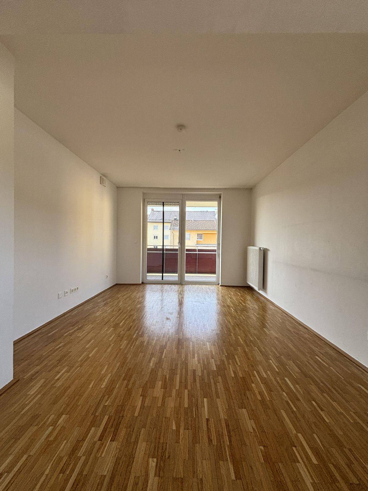 Geräumiges, leeres Zimmer mit Parkettboden und Zugang zu einem Balkon durch eine große Glastür.