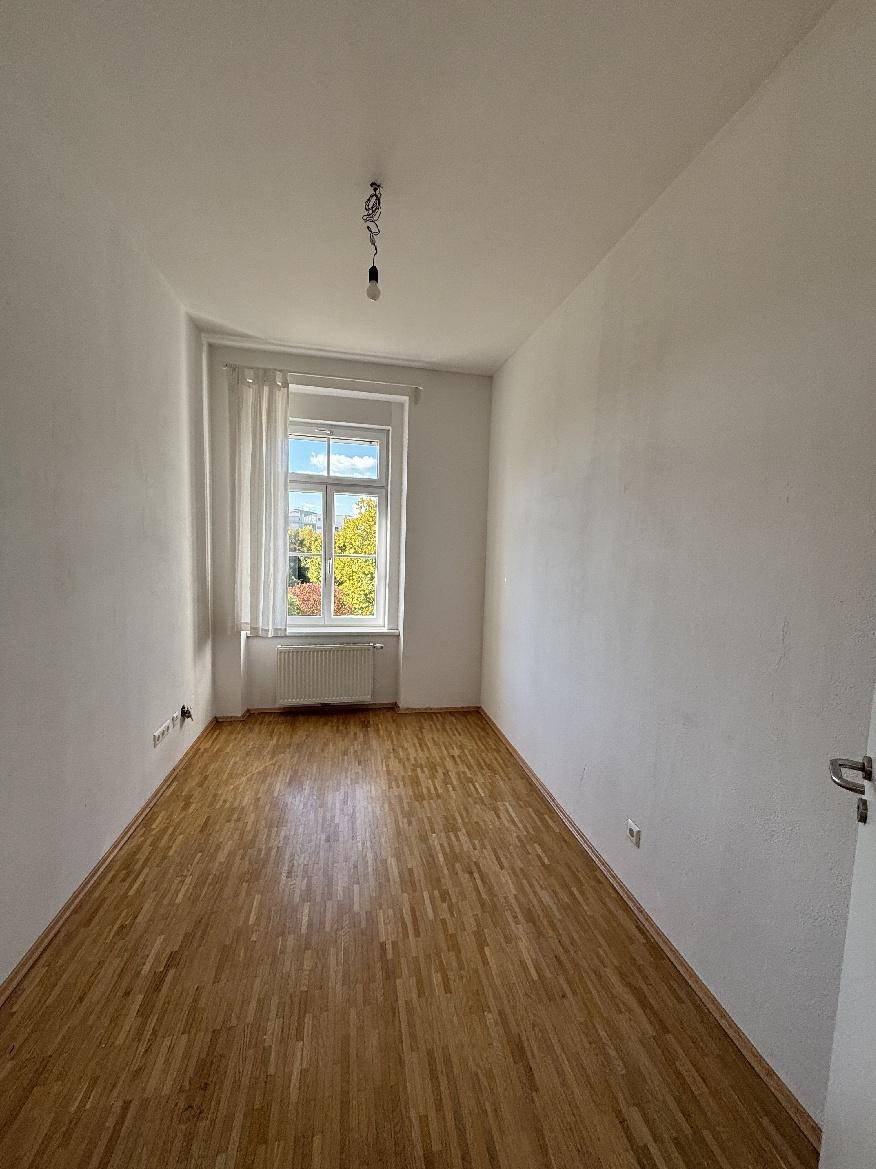 Helles Zimmer mit Parkettboden, Vorhängen und einem Fenster, das viel Tageslicht hereinlässt.