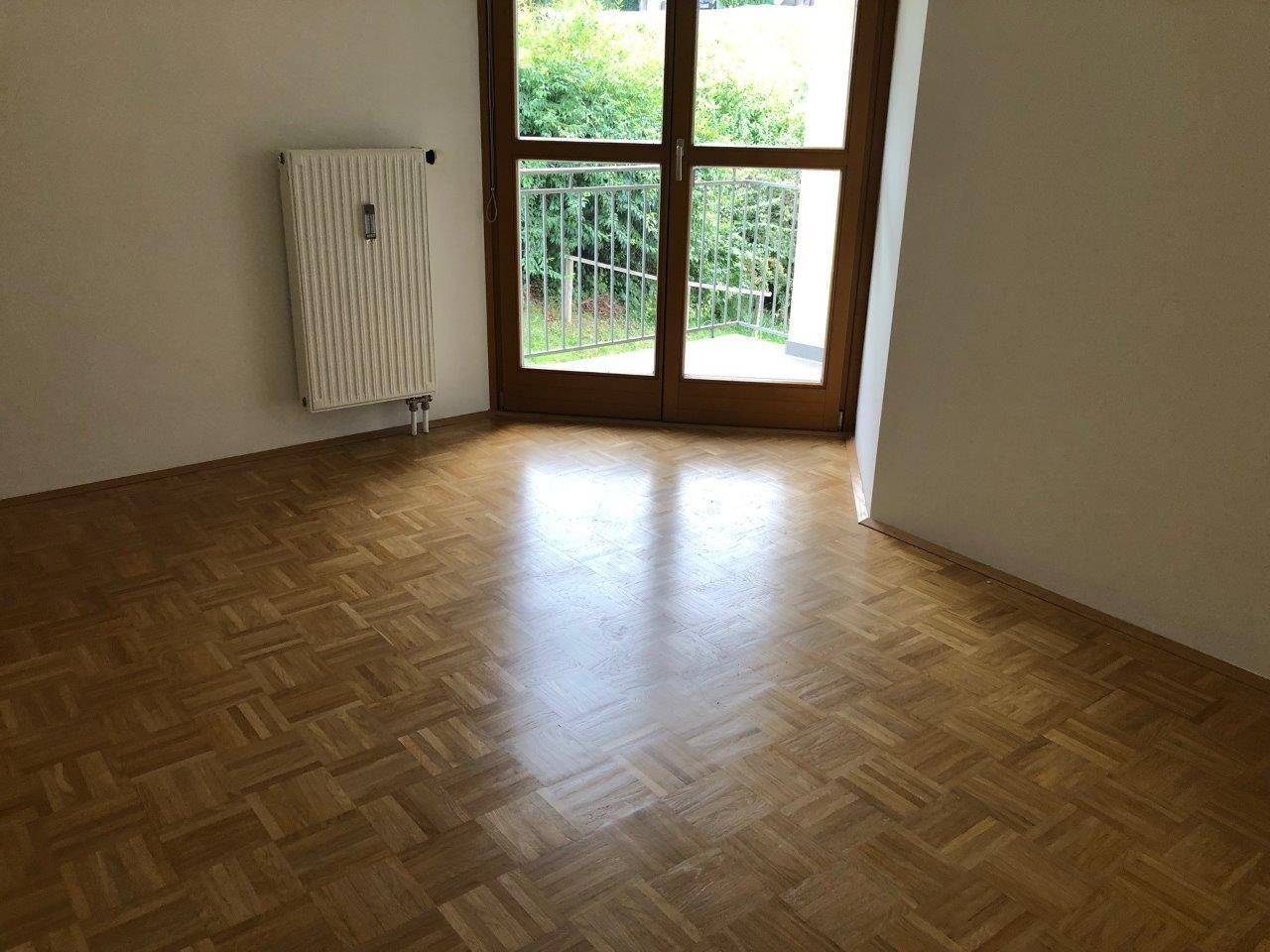 Lichtdurchfluteter Raum mit Parkettboden und großem Fenster, das Zugang zum Außenbereich bietet.