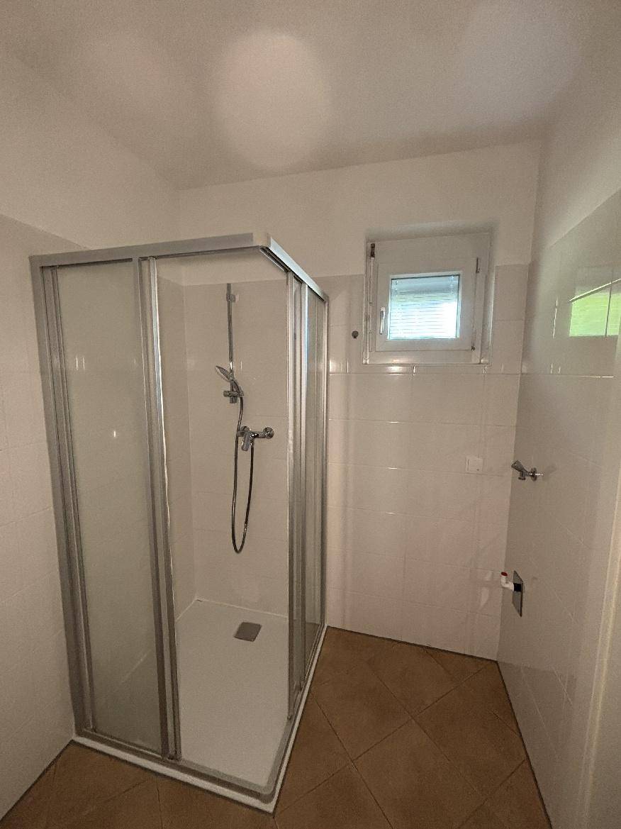 Helles Badezimmer mit Duschkabine und Fenster, weiß gefliest und mit braunem Fliesenboden.