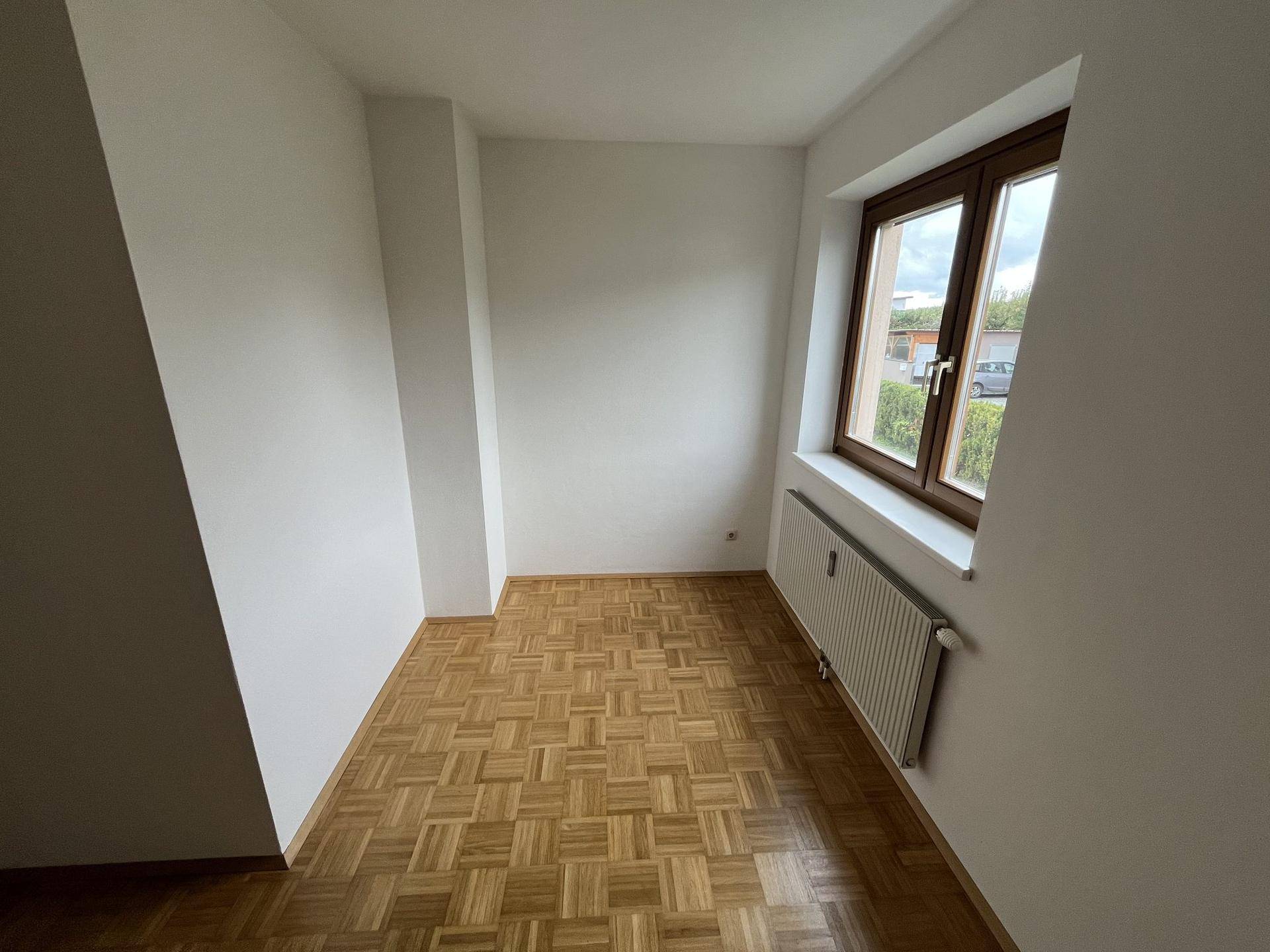 Kleineres Schlafzimmer mit Parkettboden, weißen Wänden und Fenster mit Blick ins Grüne.