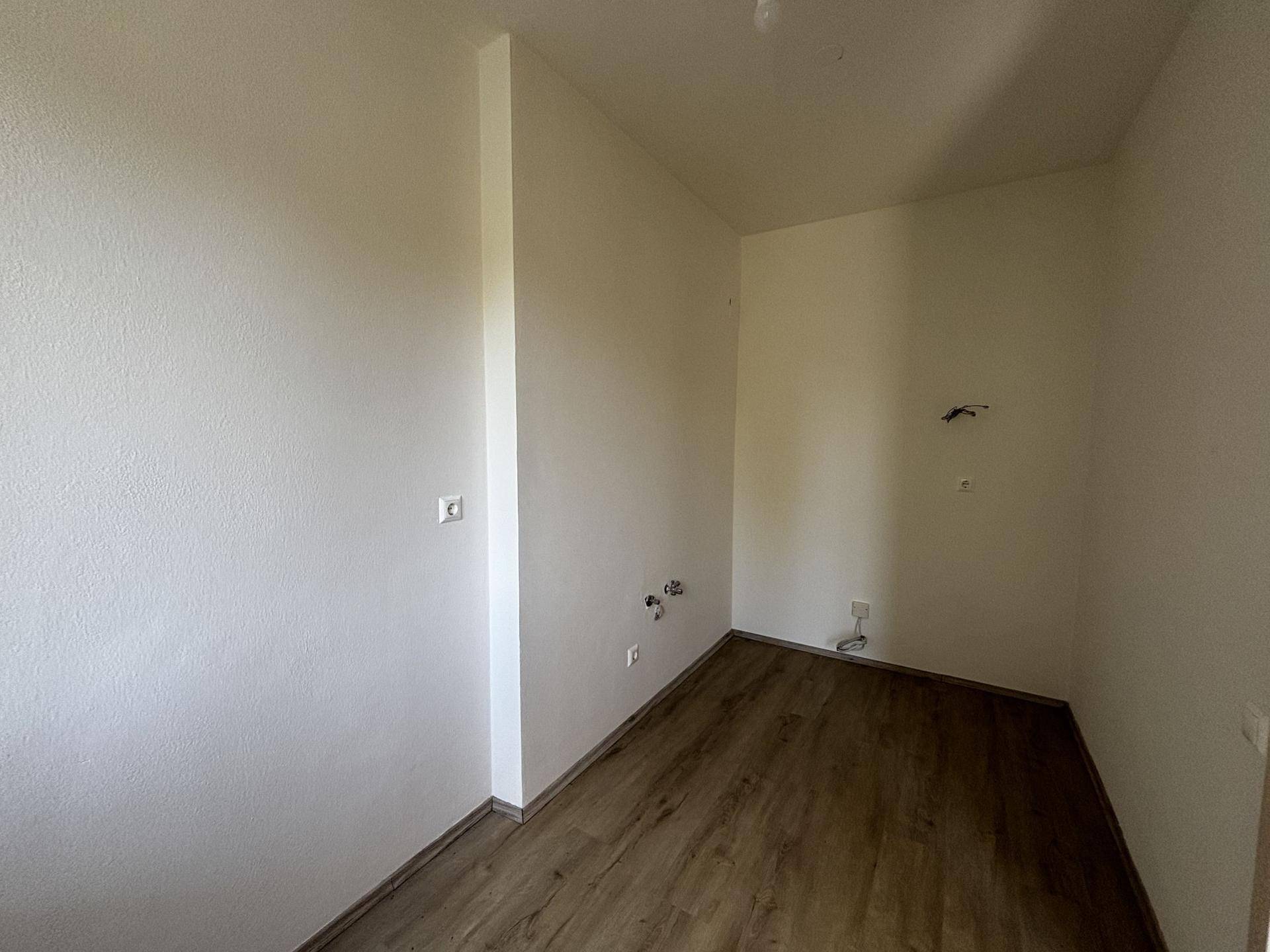 Leerer Raum mit Laminatboden und vorbereiteten Anschlüssen für eine Küche oder Waschküche.