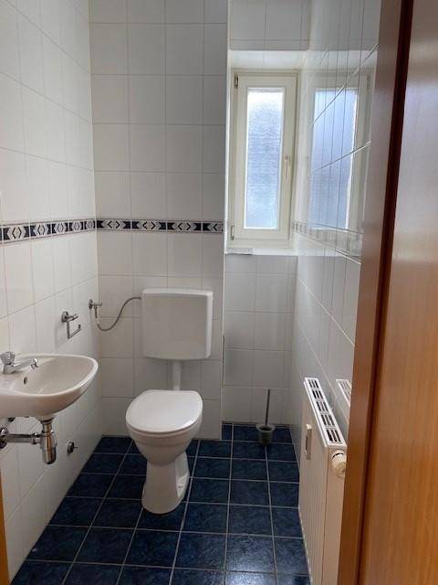 Gäste-WC mit blauen Fliesen, weißer Toilette, Waschbecken und einem Fenster für Belüftung.