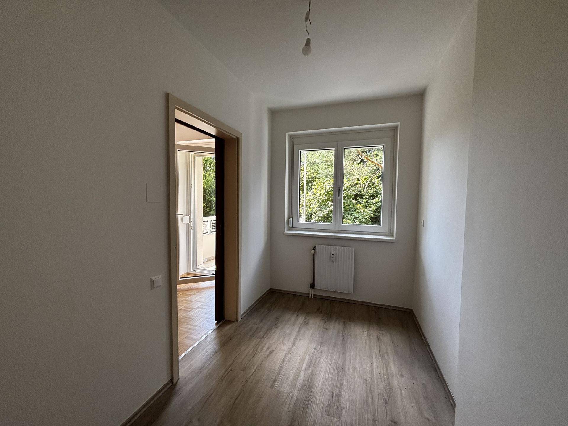 Helles Zimmer mit Laminatboden, Fenster und Durchgang zu einem weiteren Bereich der Wohnung.