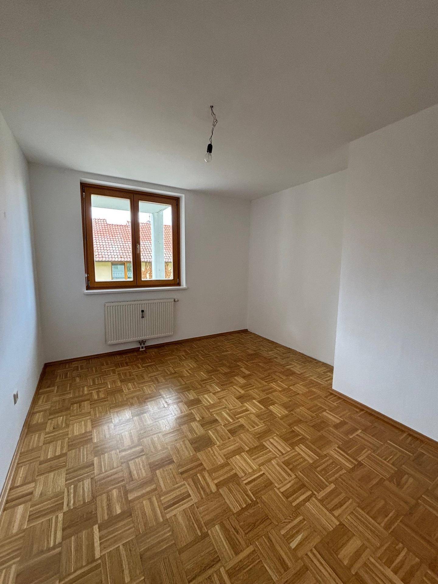 Helles Schlafzimmer mit Parkettboden, weißen Wänden und einem Fenster mit Blick ins Grüne.