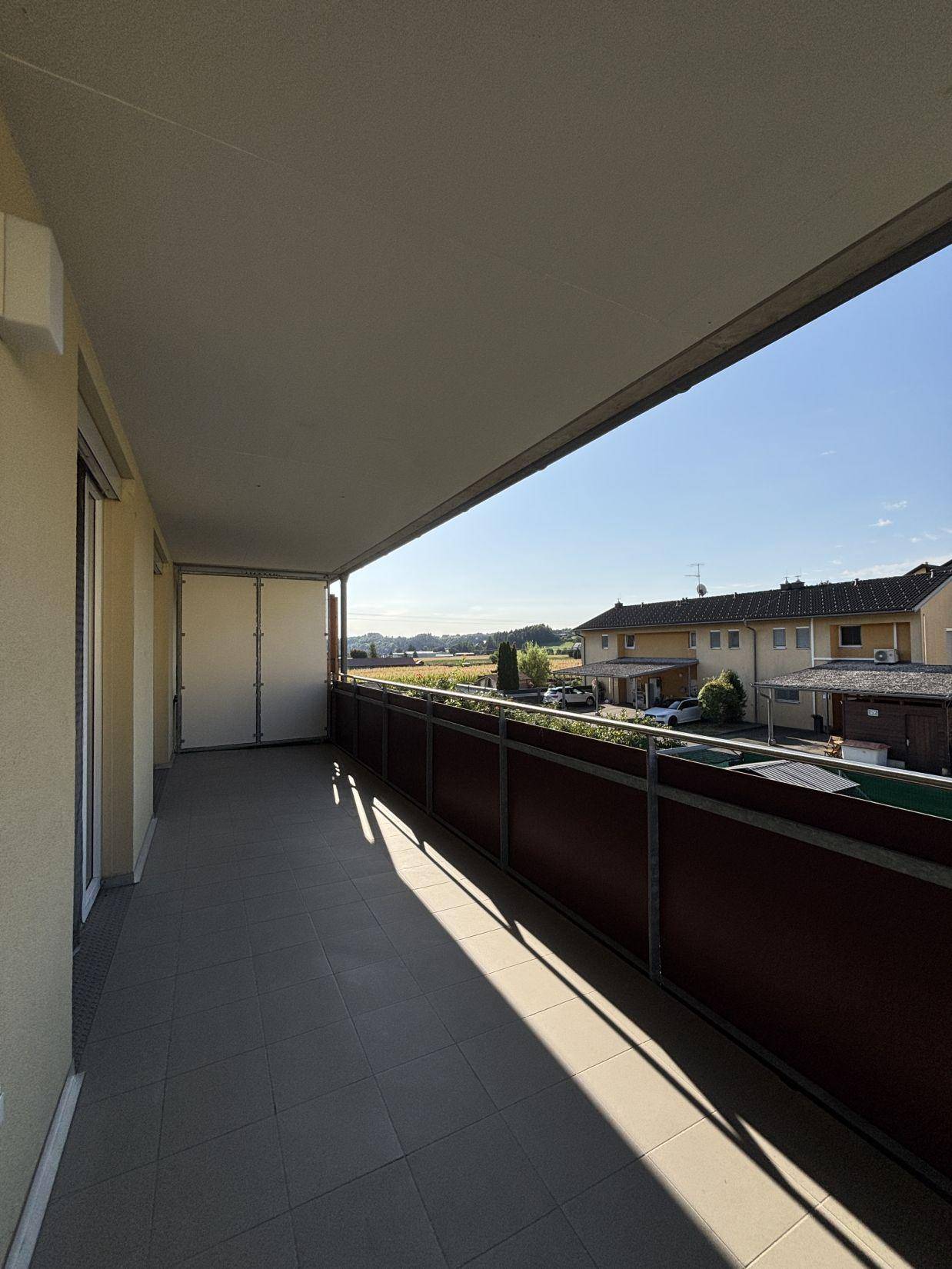 Langer, sonniger Balkon mit Fliesenboden und Blick auf die umliegende Wohnsiedlung und grüne Hügel.