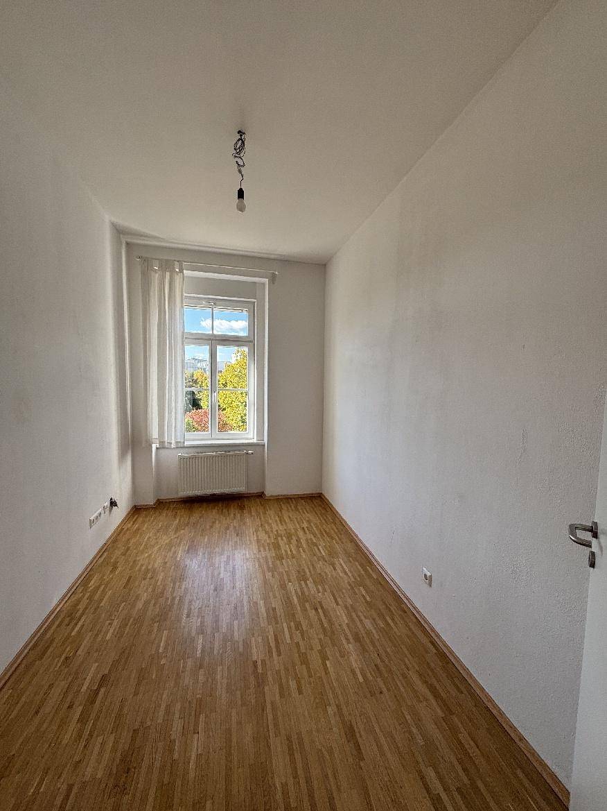 Helles Zimmer mit Parkettboden, Vorhängen und einem Fenster, das viel Tageslicht hereinlässt.