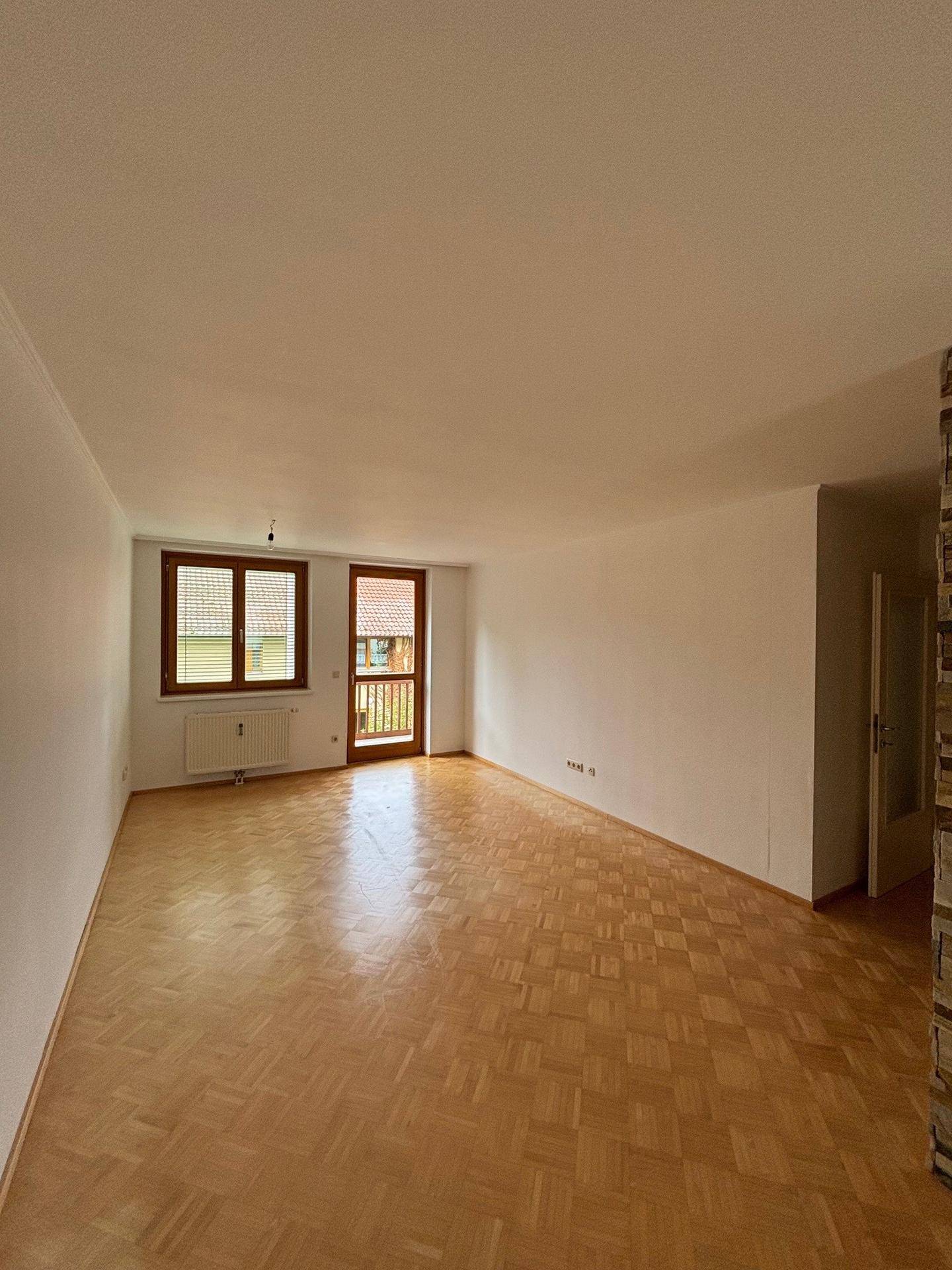 Helles Wohnzimmer mit Parkettboden, weißen Wänden und Zugang zu einem Balkon.