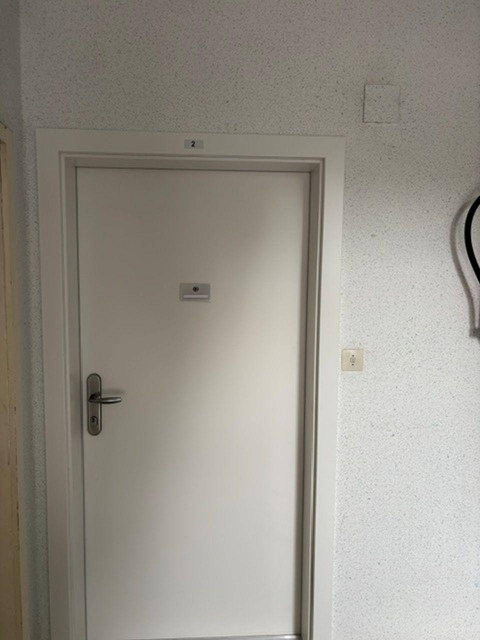 Weiße Innentür mit silbernem Griff in einem Flur mit heller, gesprenkelter Tapete und Steckdose.