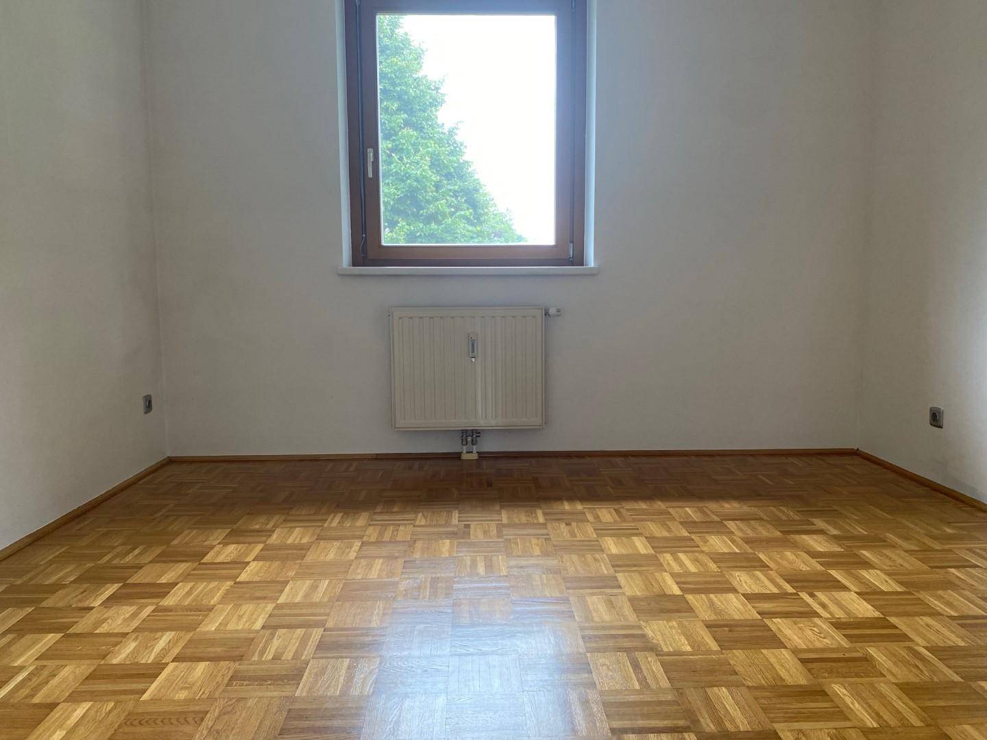 Helles Zimmer mit Parkettboden und einem großen Fenster, das viel Licht hereinlässt.