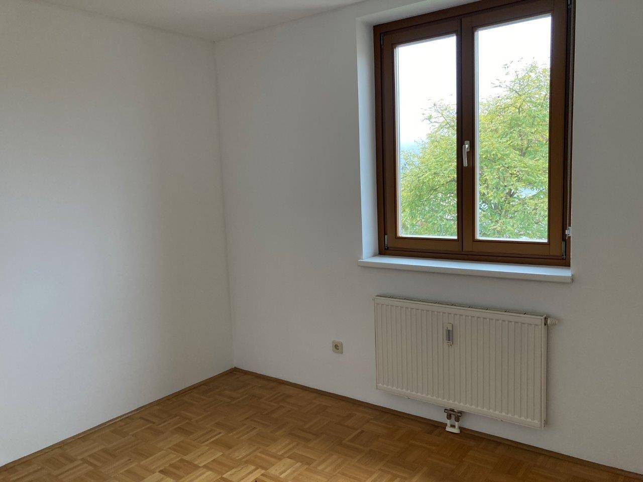 Helles Zimmer mit Parkettboden, großem Fenster und Ausblick auf Bäume.