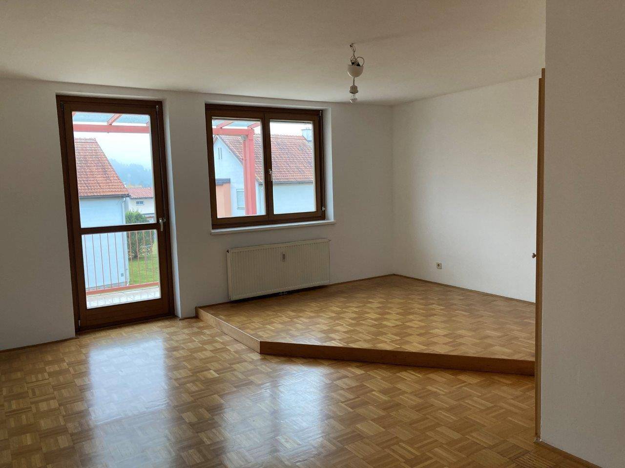 Geräumiges Wohnzimmer mit Parkettboden, erhöhter Fläche und Zugang zum Balkon.
