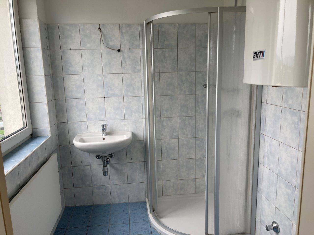 Badezimmer mit Duschkabine, Waschbecken, Fenster und hellblauen Wand- und Bodenfliesen.