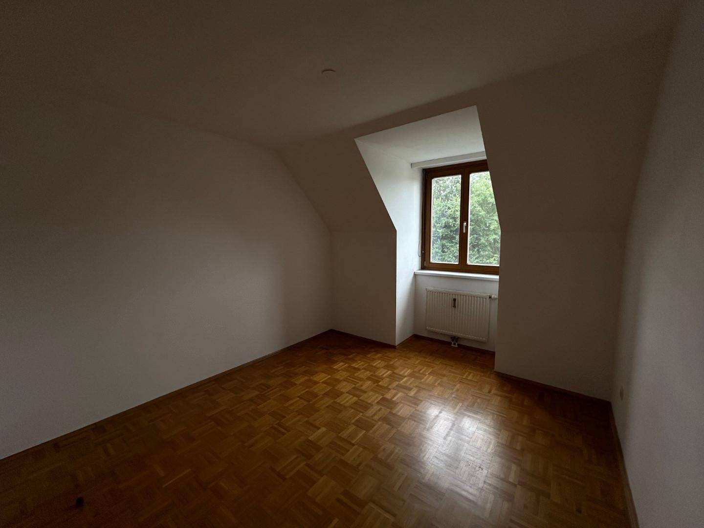 Helles Schlafzimmer mit Parkettboden, Dachschrägen und Fenster mit Blick ins Grüne.