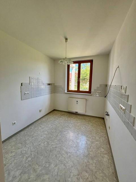 Leere Küche mit Fliesenboden, Fenster und vorbereiteten Anschlüssen für eine Einbauküche.