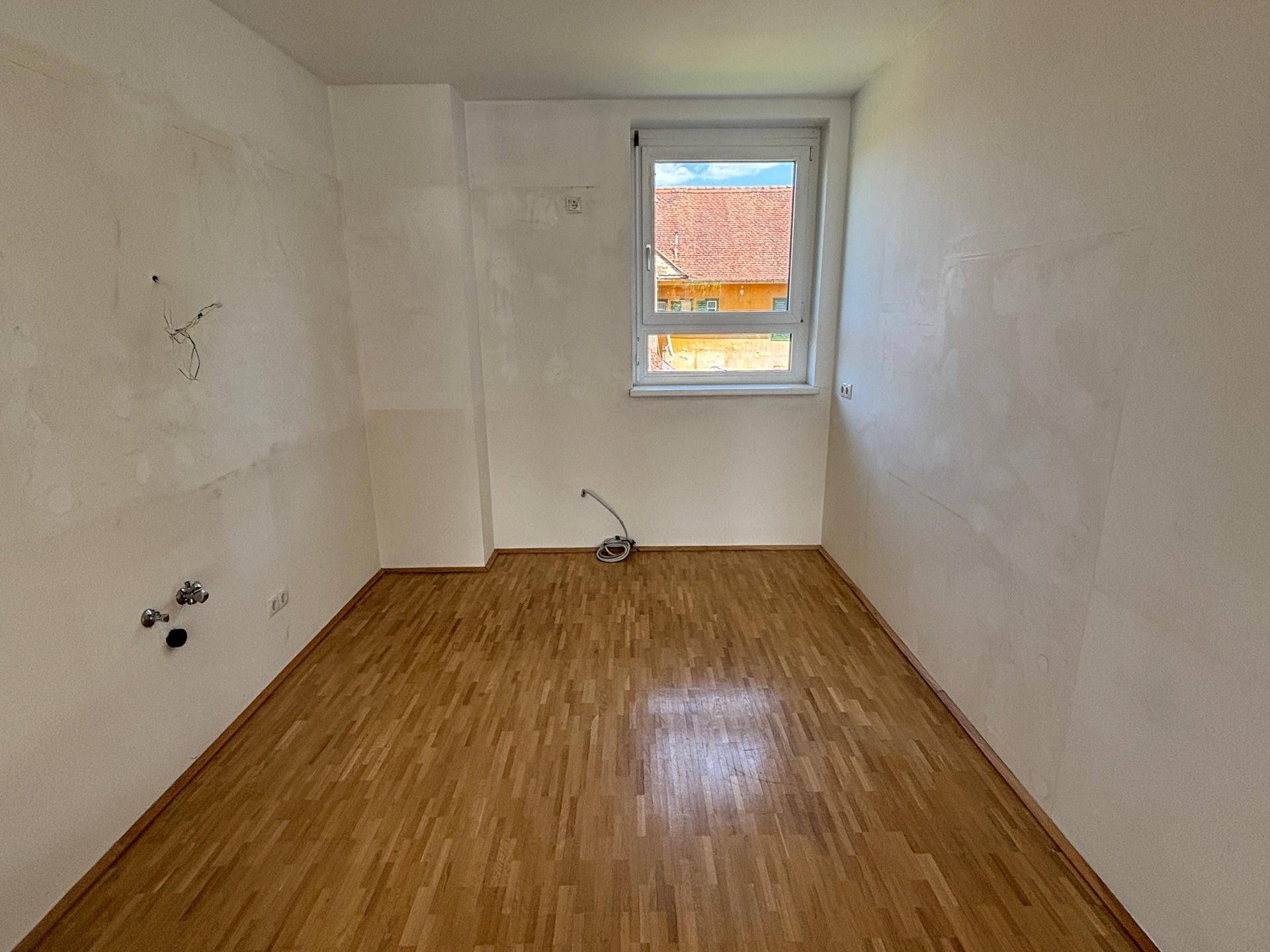 Unmöbliertes Zimmer mit Holzboden und Anschlüssen für eine mögliche Küche, bereit zur Gestaltung.