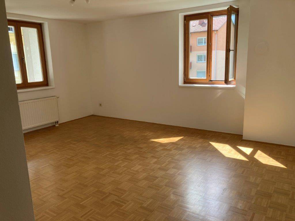 Helles Wohnzimmer mit Parkettboden und zwei großen Fenstern, die viel Tageslicht hereinlassen.