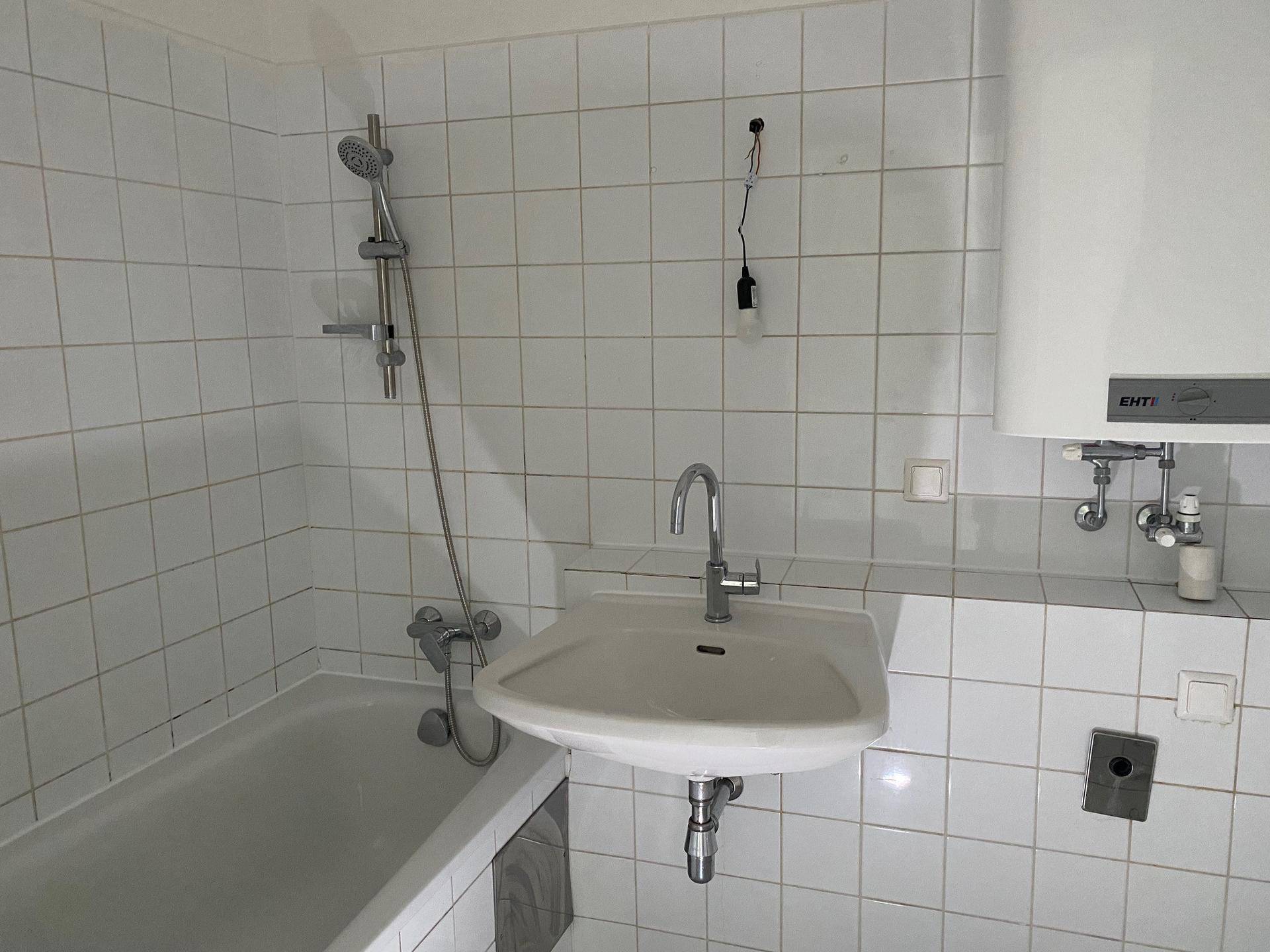 Badezimmer mit weißen Wandfliesen, Badewanne mit Duschfunktion, Waschbecken und Warmwasserboiler.