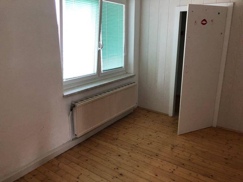 Leeres Zimmer mit Holzdielenboden, Fenster und Heizkörper, eine Tür steht offen.