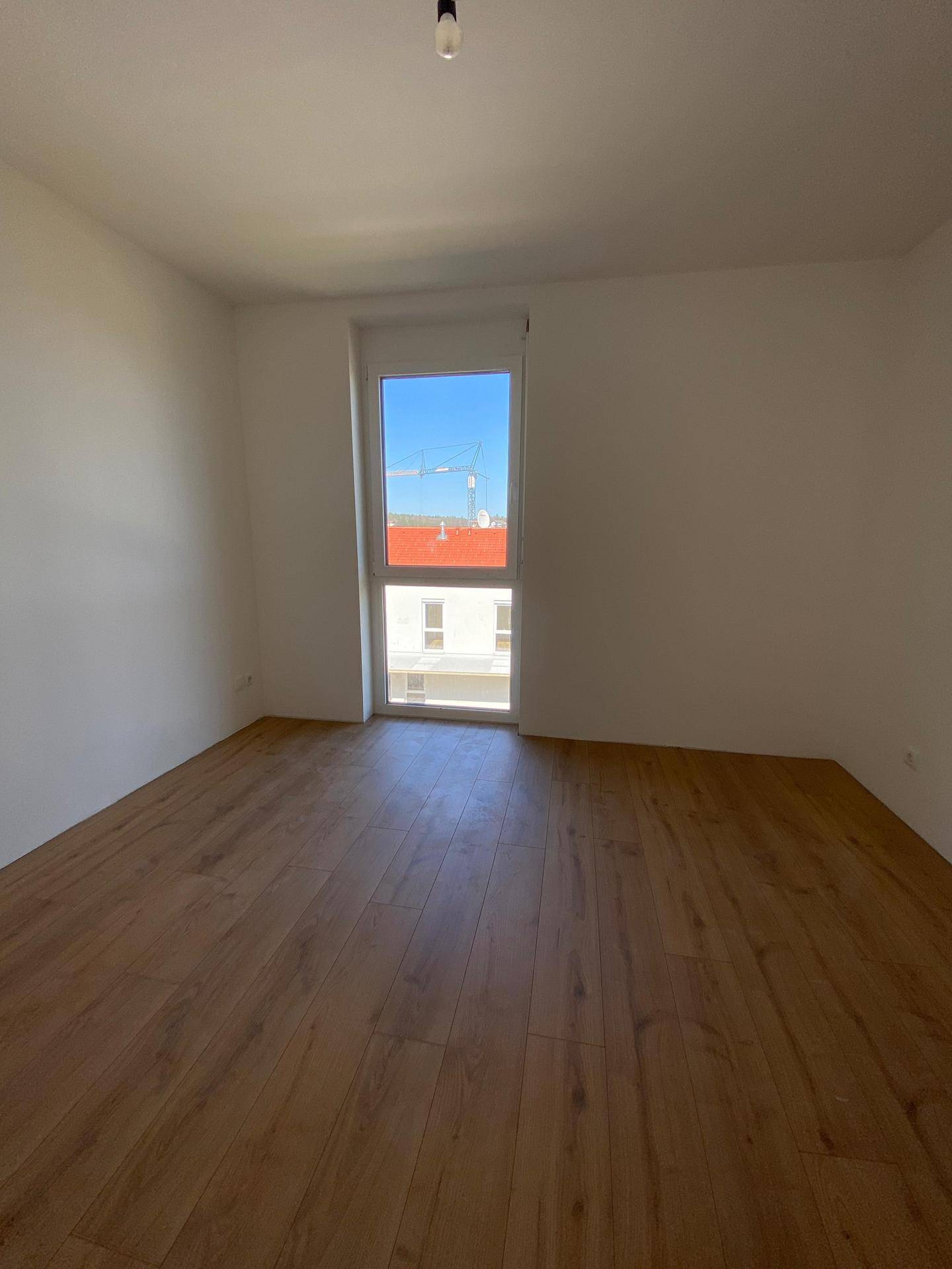 Leeres Zimmer mit Laminatboden und einem großen Fenster, das viel Licht spendet.