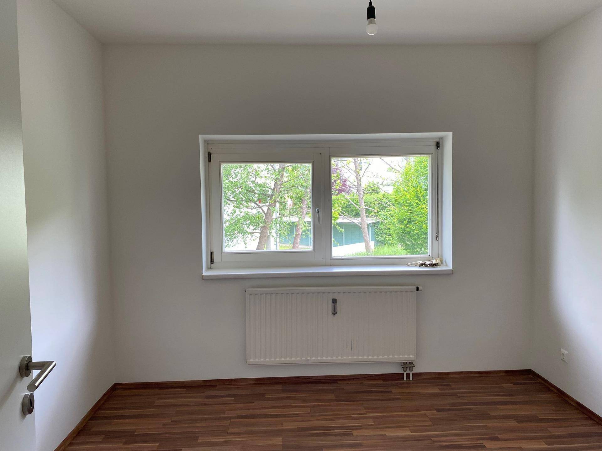 Leerer Raum mit Laminatboden, weißen Wänden und einem Doppelfenster mit Blick ins Grüne.