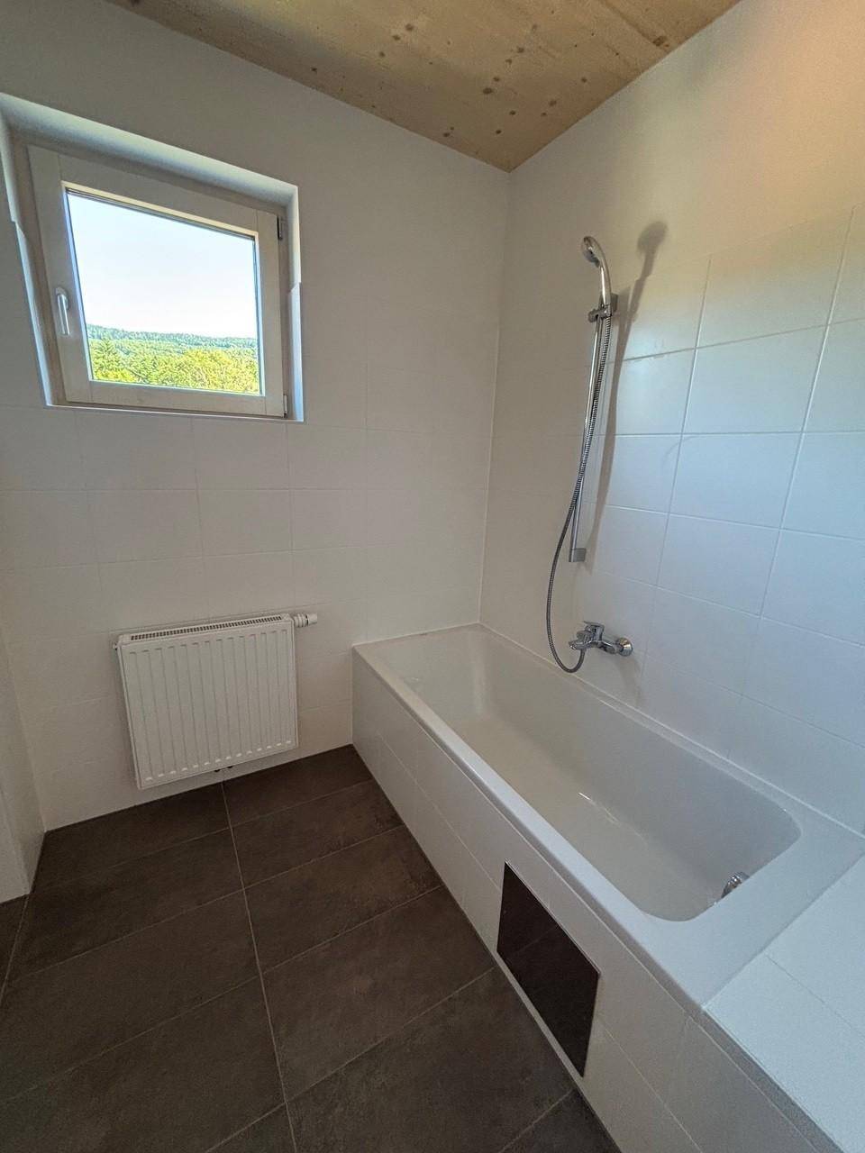 Badezimmer mit Badewanne, weißen Wandfliesen, dunklem Fliesenboden und Fenster mit Ausblick.