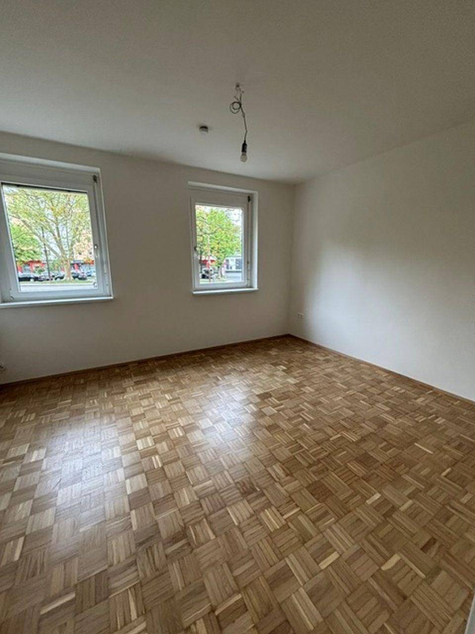Helles Schlafzimmer mit Parkettboden und zwei Fenstern, die einen Blick ins Grüne bieten.