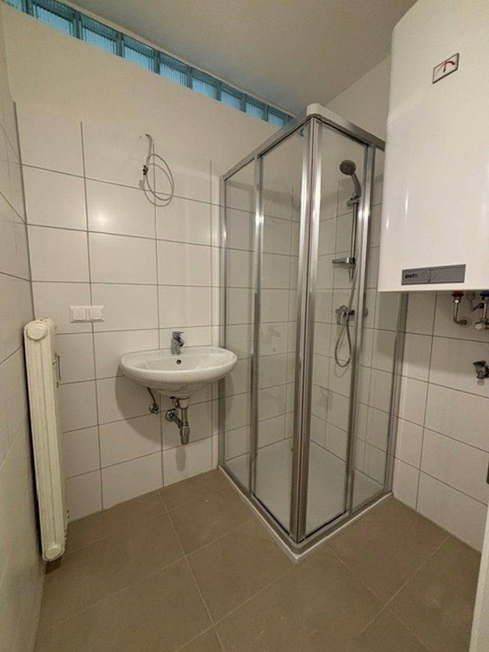 Badezimmer mit Duschkabine, Waschbecken und Boiler, ausgestattet mit weißen Fliesen und Heizkörper.