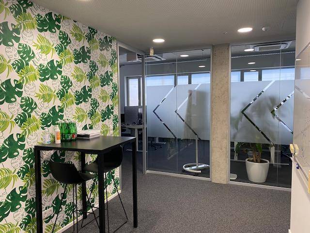 Flur mit Stehtisch und Stühlen, dekoriert mit auffälliger grüner Tapete und Glaswänden zu Büroräumen.