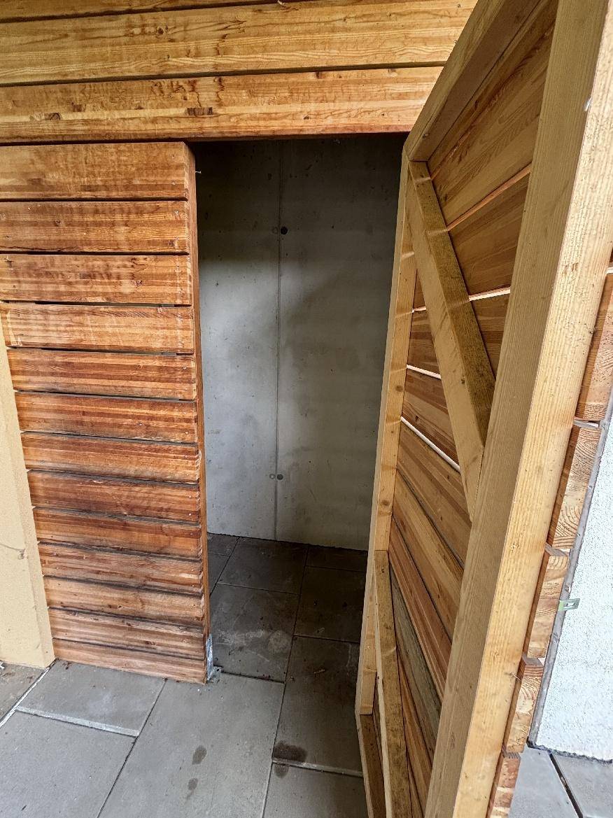 Kleiner Abstellraum mit Holzschiebetür, Betonwänden und Fliesenboden.