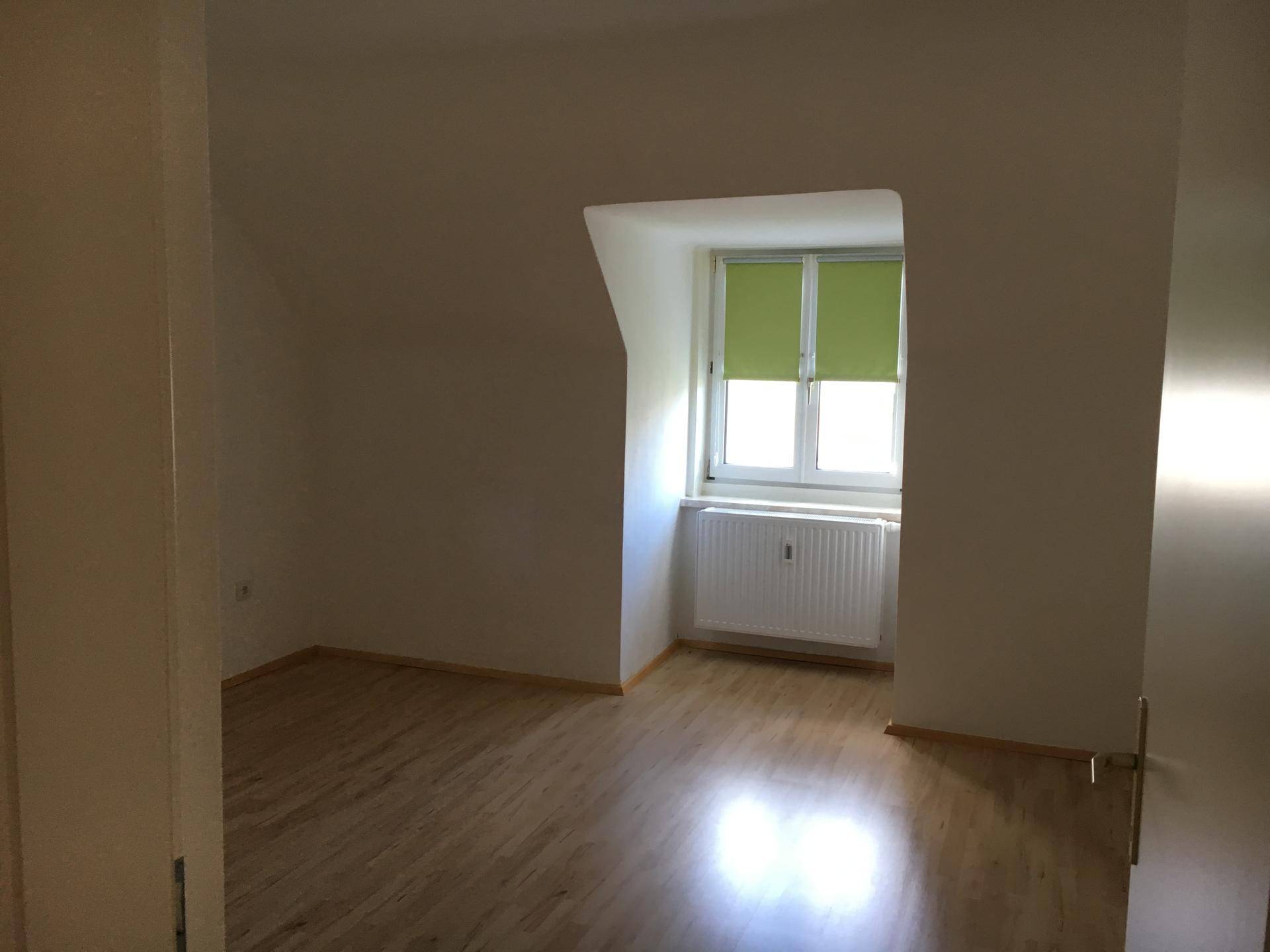 Helles Zimmer mit Holzfußboden und einem Fenster mit grünem Rollo.