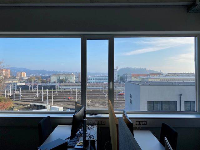 Blick aus dem Bürofenster auf ein Bahngelände und die umliegende Stadtlandschaft mit Hügeln im Hintergrund.