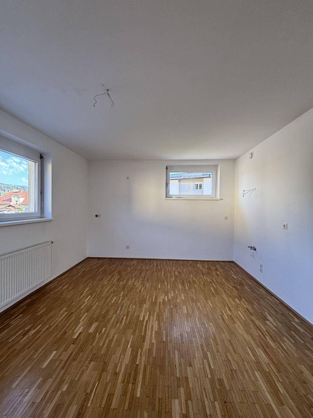 Helles, leeres Zimmer mit Parkettboden und zwei Fenstern, die einen Blick auf die Umgebung bieten.