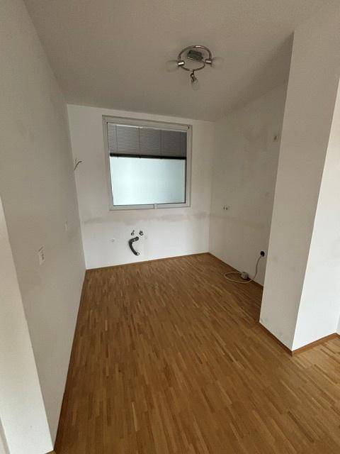 Leerer Küchenbereich mit Parkettboden, Fenster und vorbereiteten Anschlüssen für eine Küchenzeile.