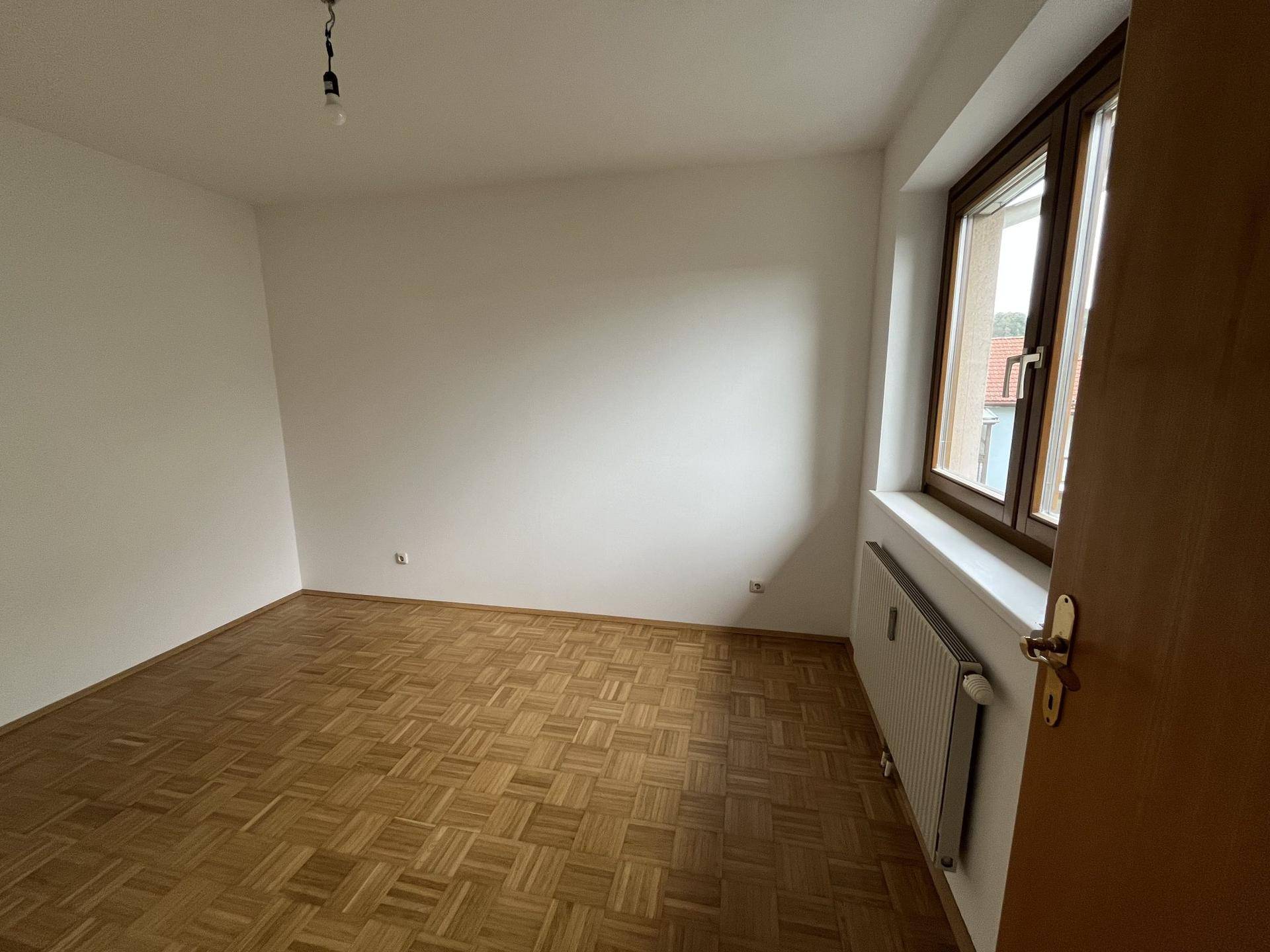 Schlafzimmer mit Parkettboden, weißen Wänden und großem Fenster mit Blick nach draußen.