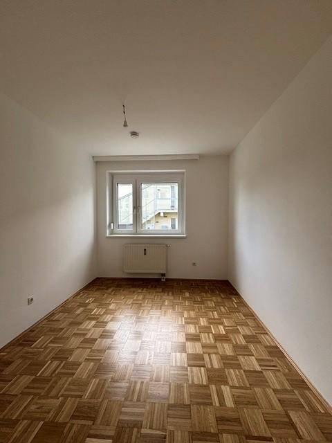 Helles Schlafzimmer mit Parkettboden und einem großen Fenster, das viel Tageslicht hereinlässt.