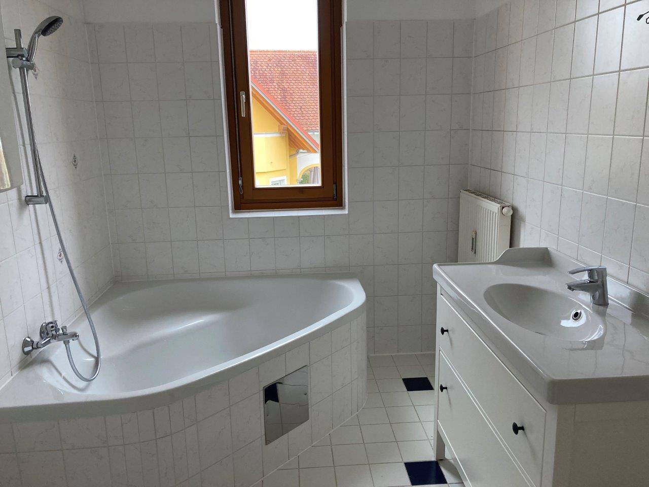 Badezimmer mit Eckbadewanne, Waschtisch, Fenster und weißen Wand- und Bodenfliesen.