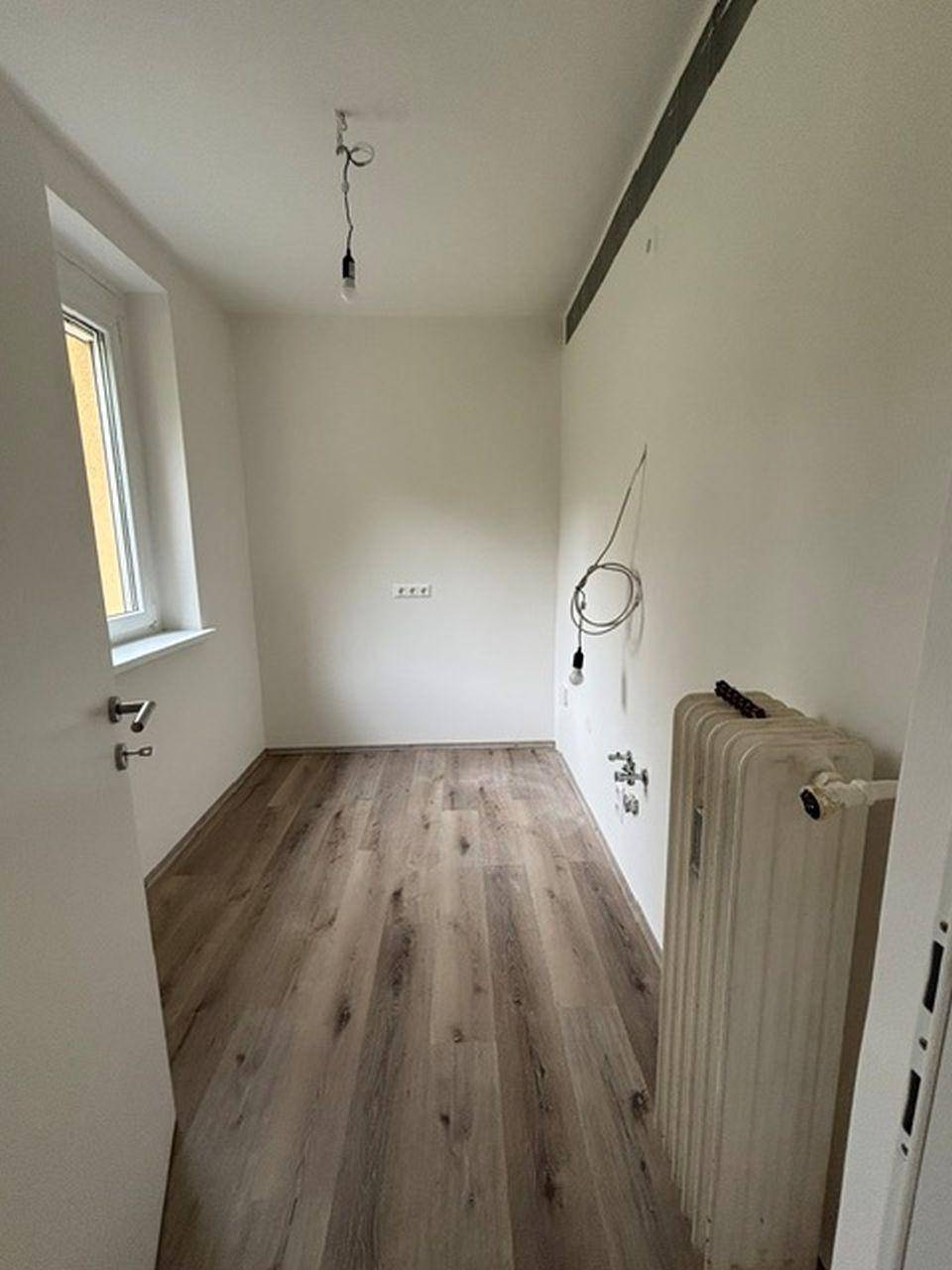 Leerer Raum mit Laminatboden, Fenster, Heizkörper und Anschlüssen für eine zukünftige Küche oder Waschküche.